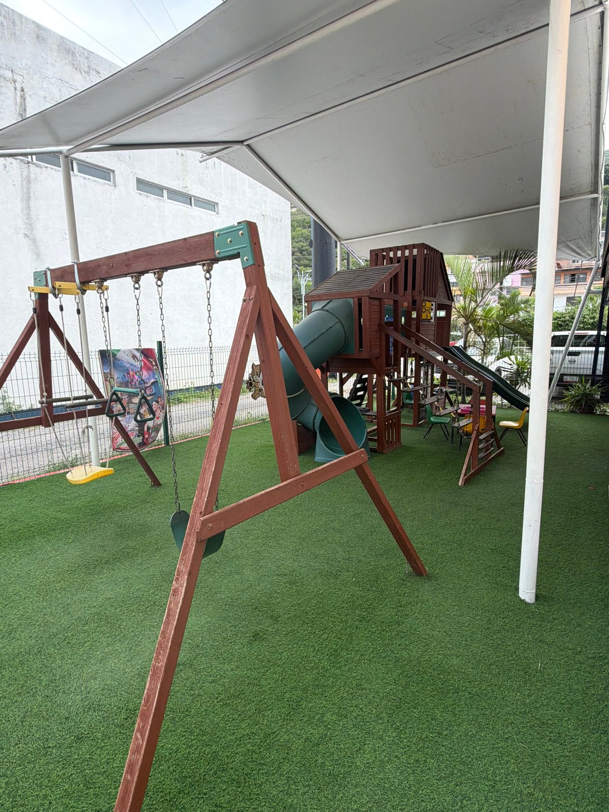 Área de juegos al aire libre con columpios de madera y estructura de escalada sobre pasto sintético